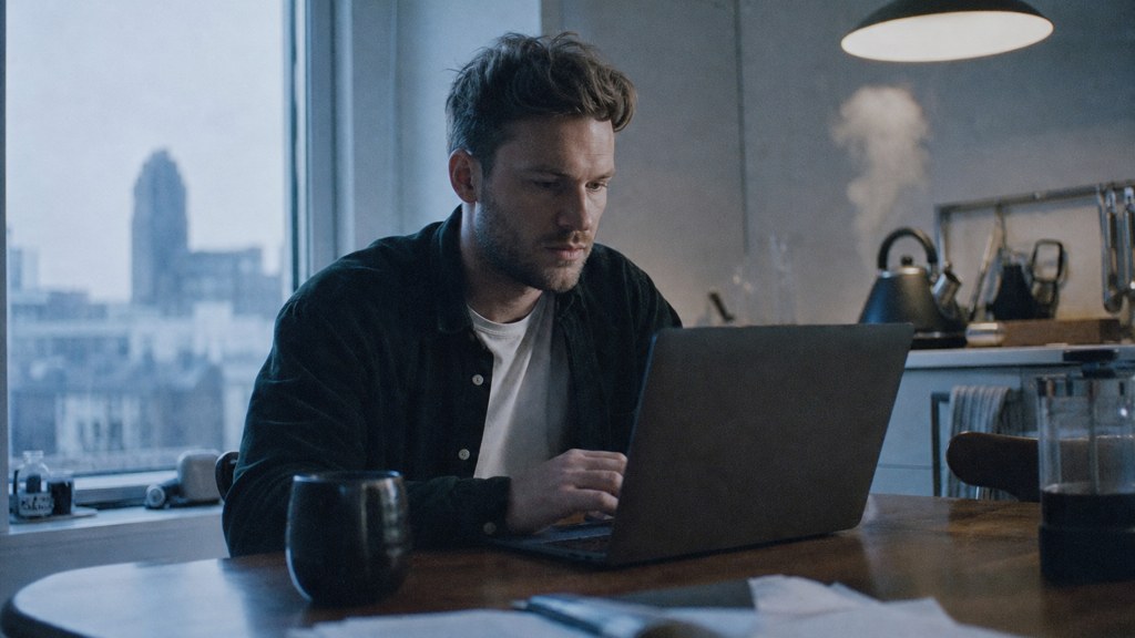 A service-business owner at a kitchen table typing a question into a laptop while a kettle steams behind, photographed in Leeds