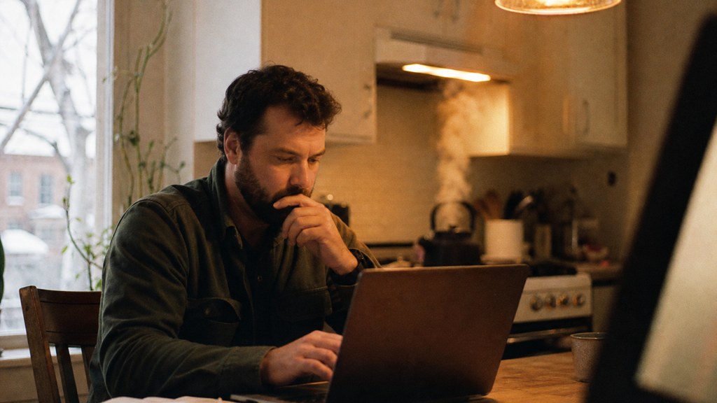 A service-business owner at a kitchen table typing a question into a laptop while a kettle steams behind, photographed in Ottawa