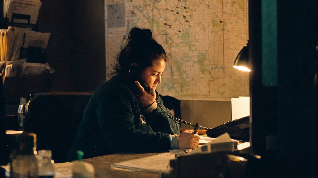 Night dispatcher at a restoration office taking an emergency call beside a service-area map