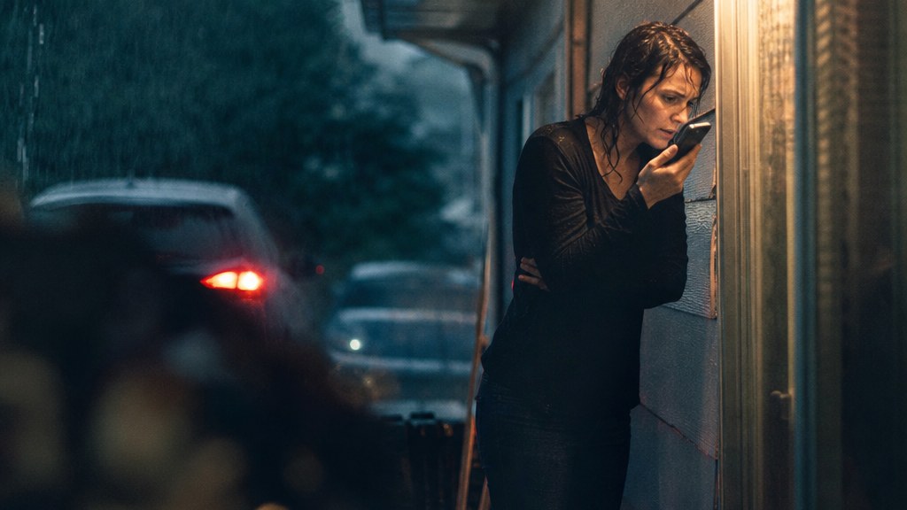 Drenched homeowner sheltering under a porch in heavy rain holding a phone close to her face