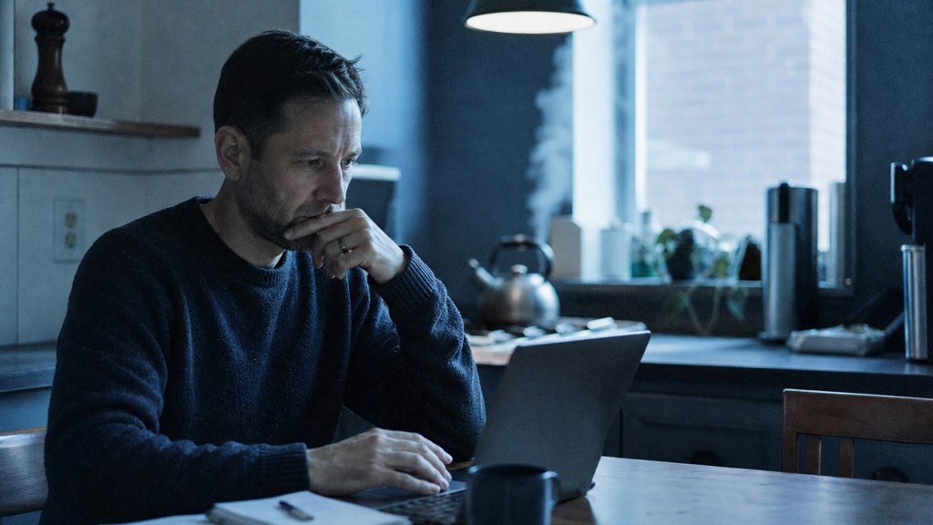A service-business owner at a kitchen table typing a question into a laptop while a kettle steams behind, photographed in Philadelphia