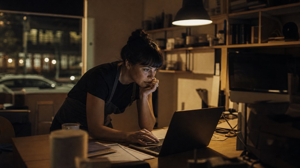 A small-business owner in an apron leaning over a laptop on a cluttered back-office desk late in the day, photographed in Adelaide