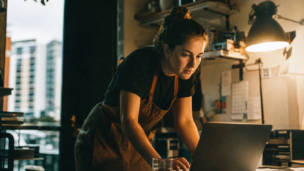A small-business owner in an apron leaning over a laptop on a cluttered back-office desk late in the day, photographed in Adelaide