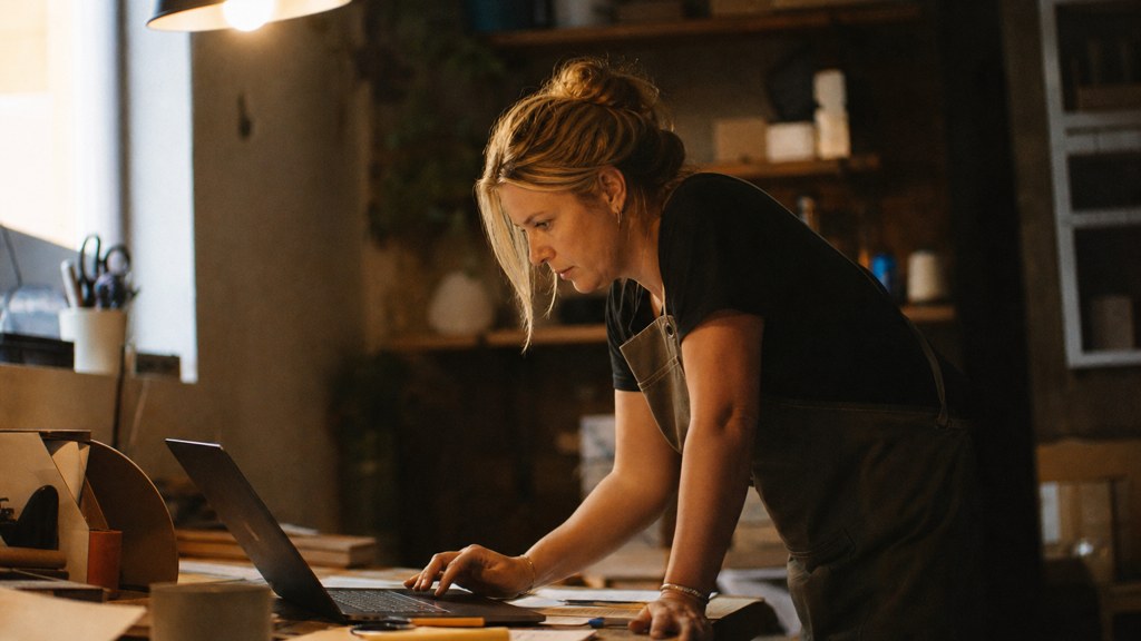 A small-business owner in an apron leaning over a laptop on a cluttered back-office desk late in the day, photographed in Adelaide