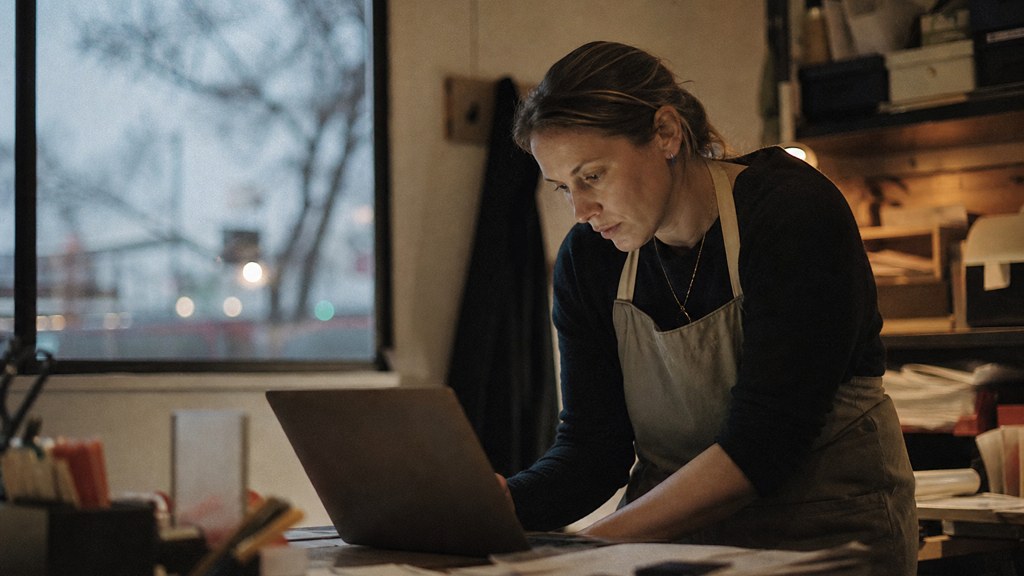A small-business owner in an apron leaning over a laptop on a cluttered back-office desk late in the day, photographed in Adelaide