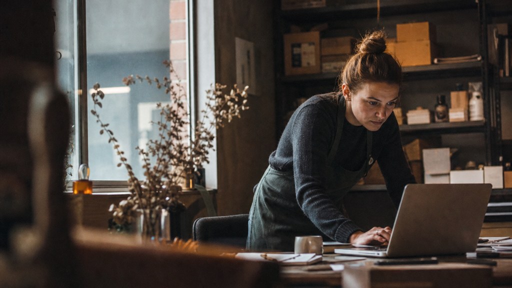 A small-business owner in an apron leaning over a laptop on a cluttered back-office desk late in the day, photographed in Adelaide