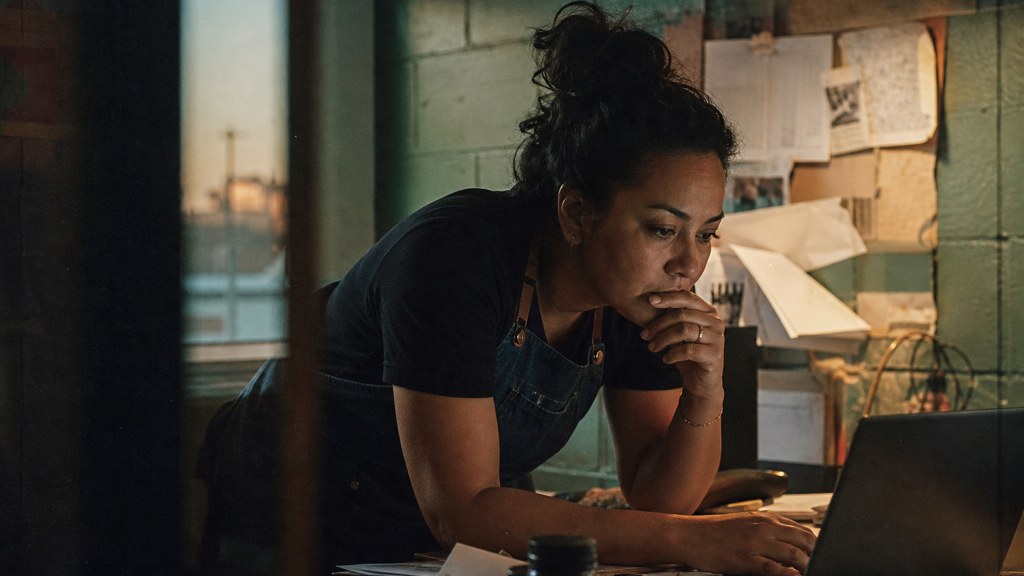 A small-business owner in an apron leaning over a laptop on a cluttered back-office desk late in the day, photographed in Albuquerque
