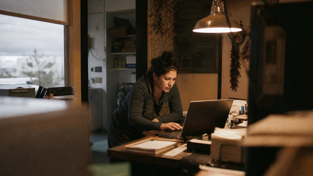 A small-business owner in an apron leaning over a laptop on a cluttered back-office desk late in the day, photographed in Albuquerque