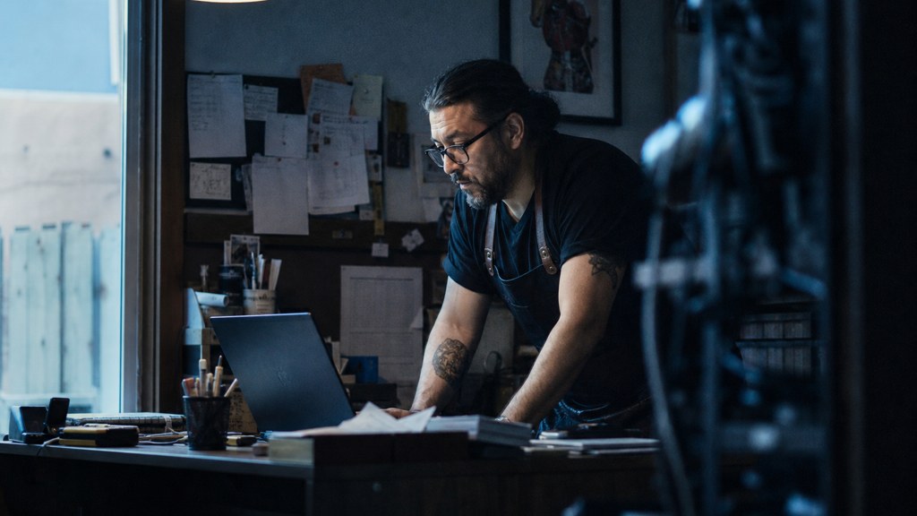 A small-business owner in an apron leaning over a laptop on a cluttered back-office desk late in the day, photographed in Albuquerque