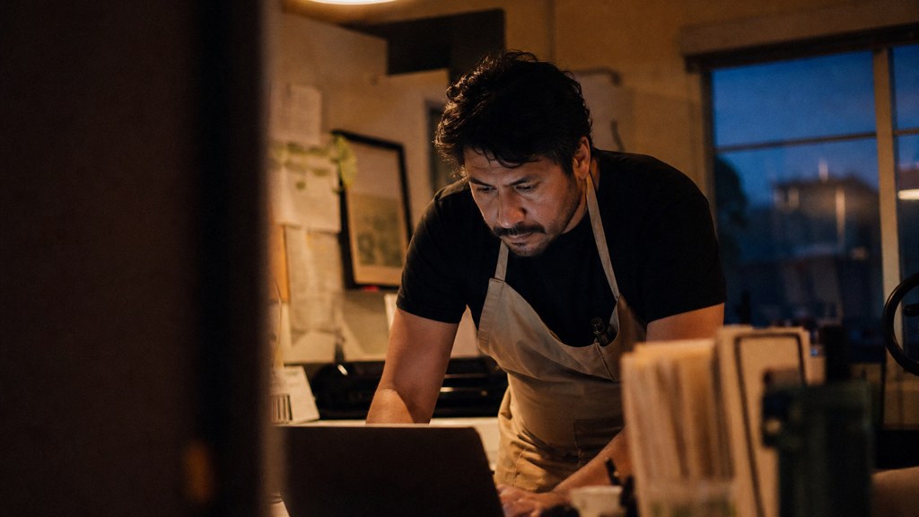 A small-business owner in an apron leaning over a laptop on a cluttered back-office desk late in the day, photographed in Albuquerque