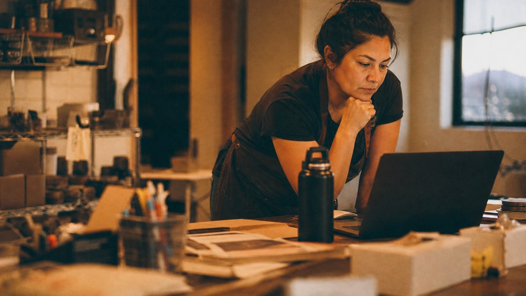 A small-business owner in an apron leaning over a laptop on a cluttered back-office desk late in the day, photographed in Albuquerque