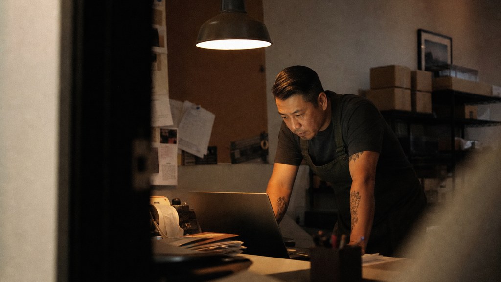 A small-business owner in an apron leaning over a laptop on a cluttered back-office desk late in the day, photographed in Anaheim