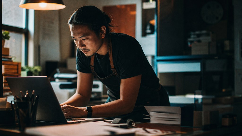 A small-business owner in an apron leaning over a laptop on a cluttered back-office desk late in the day, photographed in Anaheim