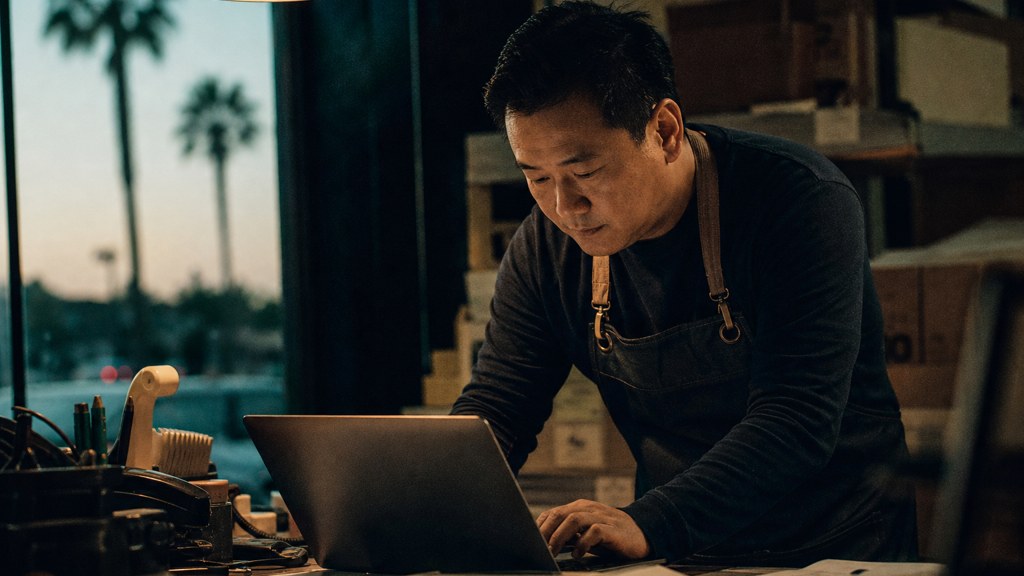 A small-business owner in an apron leaning over a laptop on a cluttered back-office desk late in the day, photographed in Anaheim