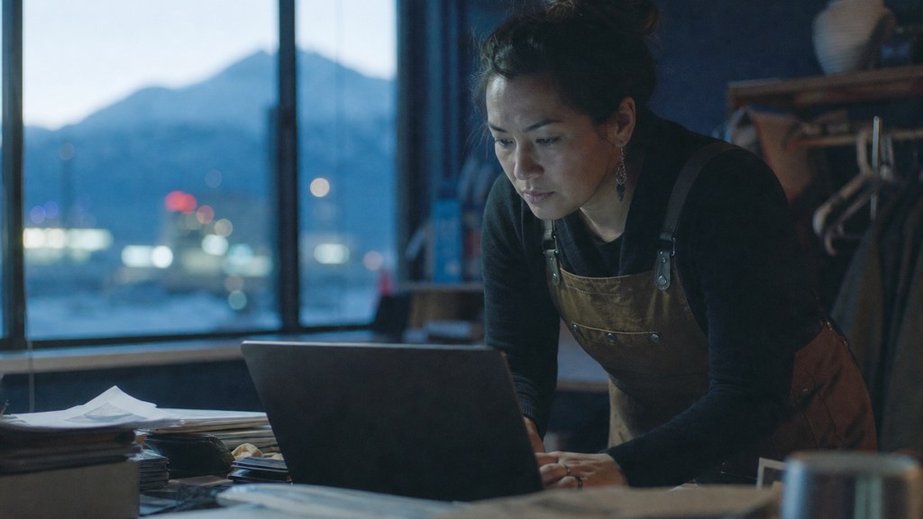 A small-business owner in an apron leaning over a laptop on a cluttered back-office desk late in the day, photographed in Anchorage