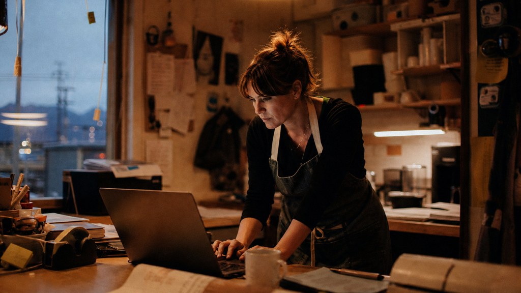 A small-business owner in an apron leaning over a laptop on a cluttered back-office desk late in the day, photographed in Anchorage