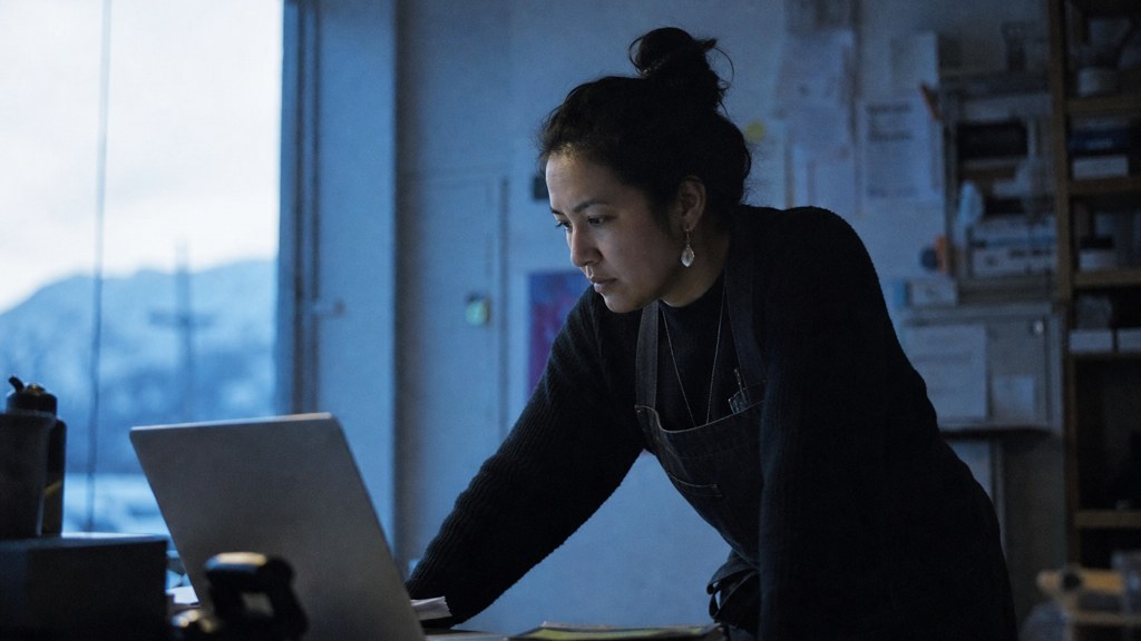 A small-business owner in an apron leaning over a laptop on a cluttered back-office desk late in the day, photographed in Anchorage