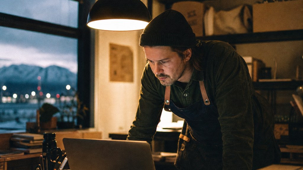 A small-business owner in an apron leaning over a laptop on a cluttered back-office desk late in the day, photographed in Anchorage