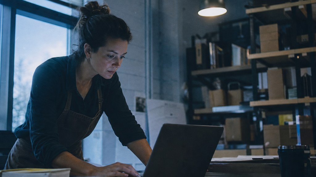 A small-business owner in an apron leaning over a laptop on a cluttered back-office desk late in the day, photographed in Arlington