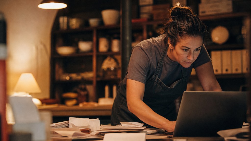 A small-business owner in an apron leaning over a laptop on a cluttered back-office desk late in the day, photographed in Arlington