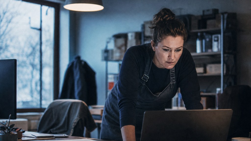 A small-business owner in an apron leaning over a laptop on a cluttered back-office desk late in the day, photographed in Arlington