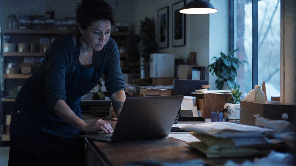 A small-business owner in an apron leaning over a laptop on a cluttered back-office desk late in the day, photographed in Arlington