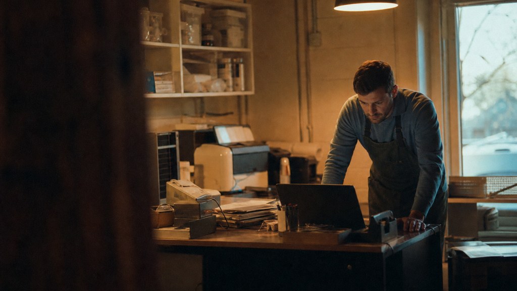 A small-business owner in an apron leaning over a laptop on a cluttered back-office desk late in the day, photographed in Arlington