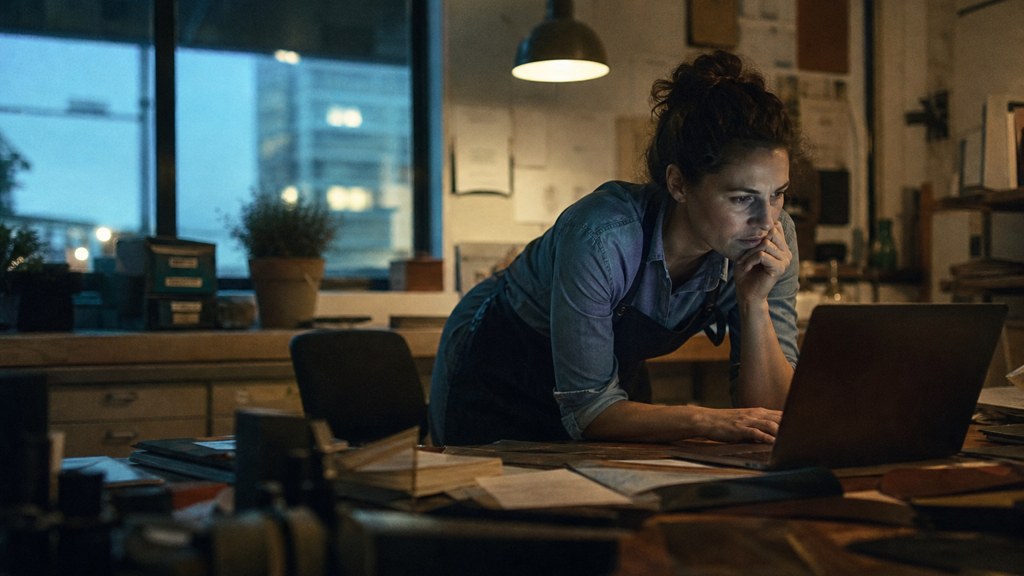 A small-business owner in an apron leaning over a laptop on a cluttered back-office desk late in the day, photographed in Arlington