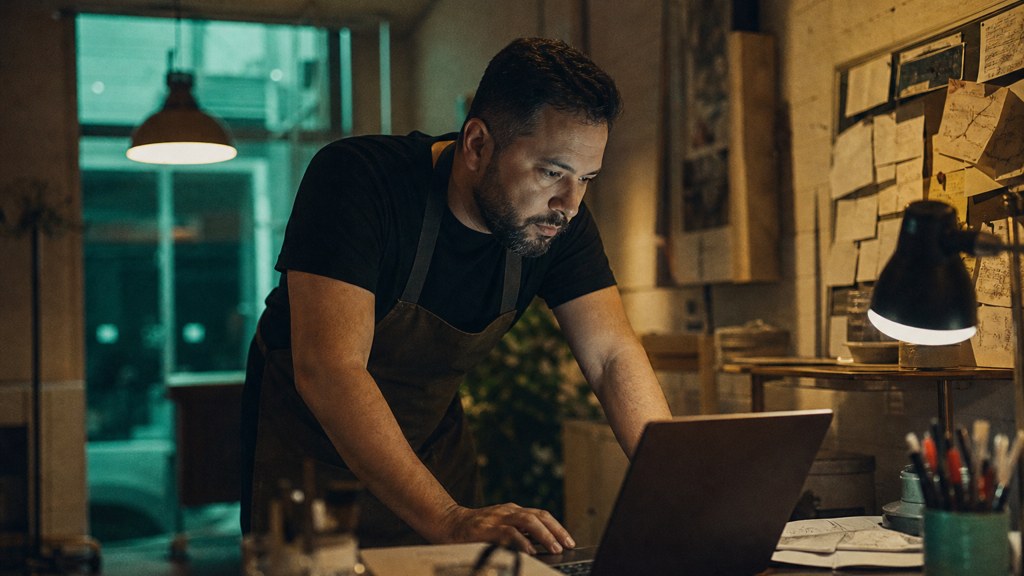 A small-business owner in an apron leaning over a laptop on a cluttered back-office desk late in the day, photographed in Arlington