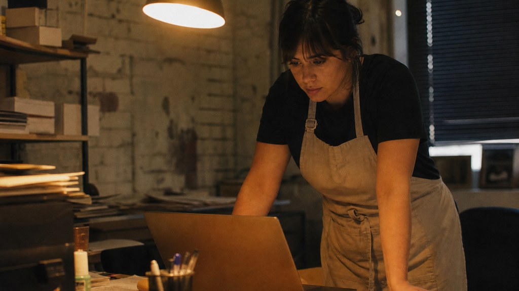 A small-business owner in an apron leaning over a laptop on a cluttered back-office desk late in the day, photographed in Arlington
