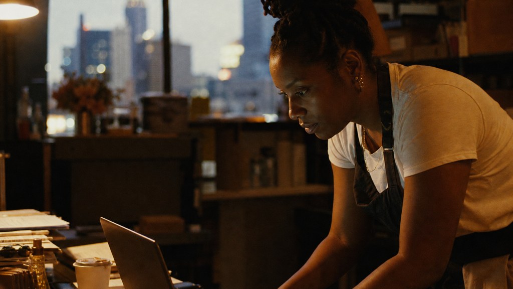 A small-business owner in an apron leaning over a laptop on a cluttered back-office desk late in the day, photographed in Atlanta