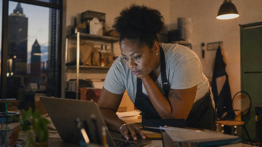 A small-business owner in an apron leaning over a laptop on a cluttered back-office desk late in the day, photographed in Atlanta