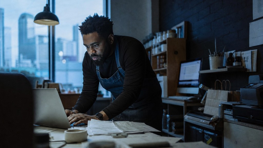 A small-business owner in an apron leaning over a laptop on a cluttered back-office desk late in the day, photographed in Atlanta
