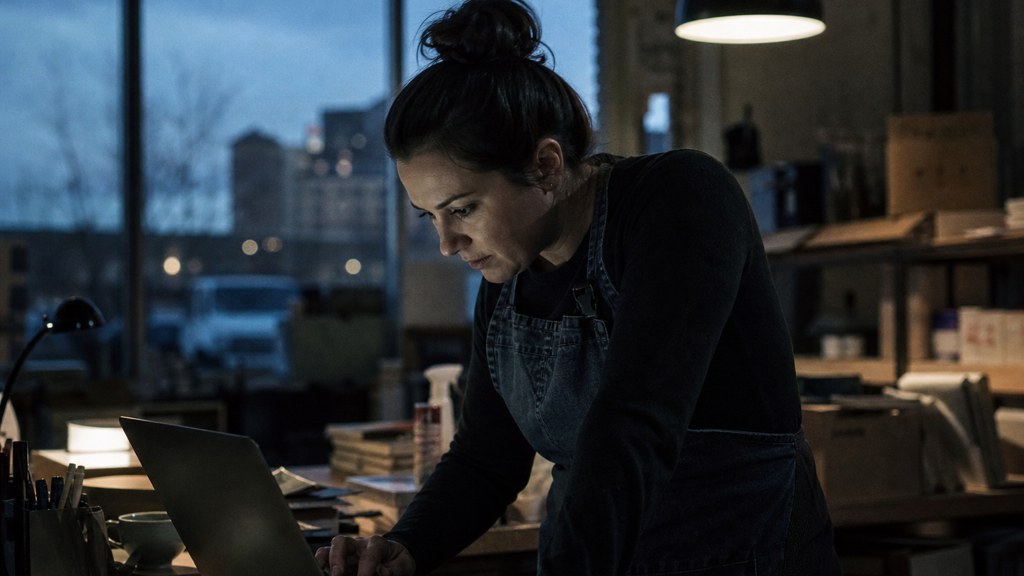 A small-business owner in an apron leaning over a laptop on a cluttered back-office desk late in the day, photographed in Aurora