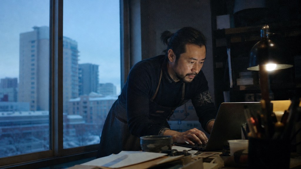 A small-business owner in an apron leaning over a laptop on a cluttered back-office desk late in the day, photographed in Aurora