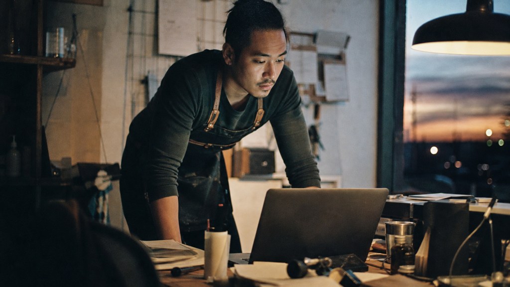 A small-business owner in an apron leaning over a laptop on a cluttered back-office desk late in the day, photographed in Aurora