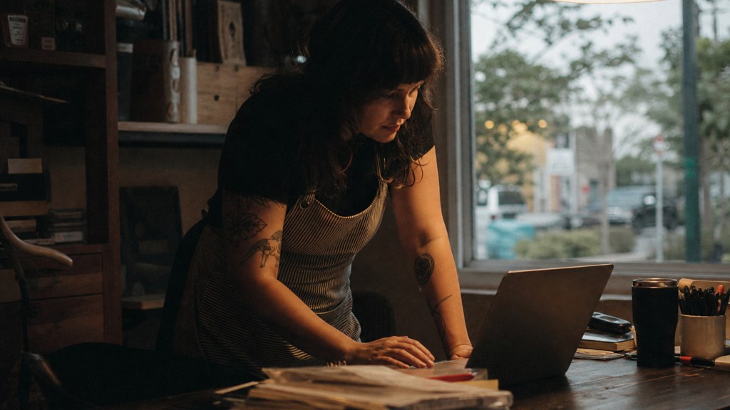 A small-business owner in an apron leaning over a laptop on a cluttered back-office desk late in the day, photographed in Austin