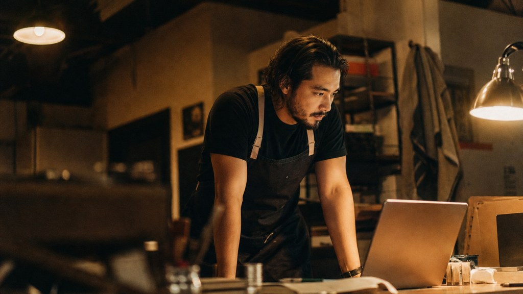 A small-business owner in an apron leaning over a laptop on a cluttered back-office desk late in the day, photographed in Austin