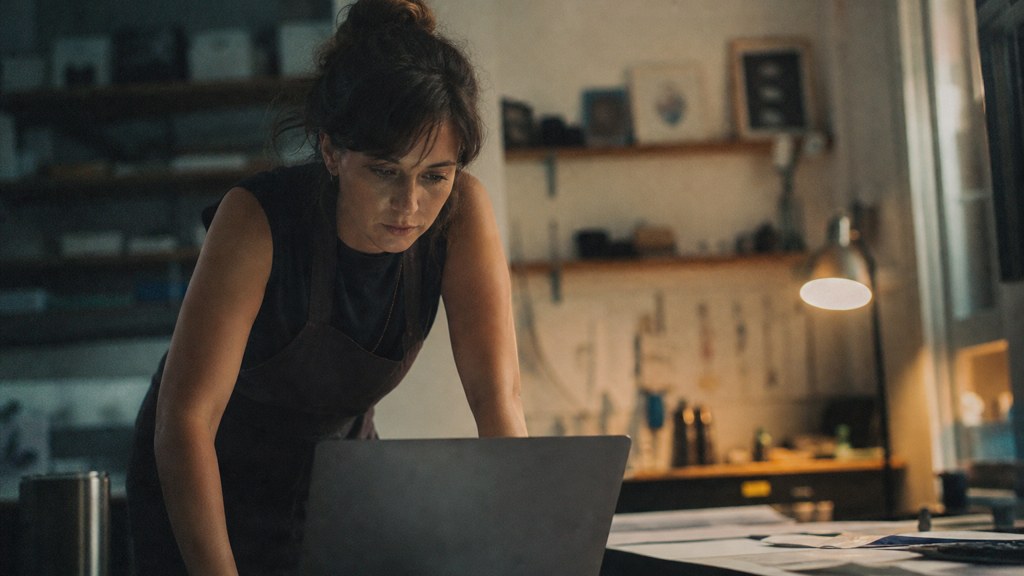 A small-business owner in an apron leaning over a laptop on a cluttered back-office desk late in the day, photographed in Austin