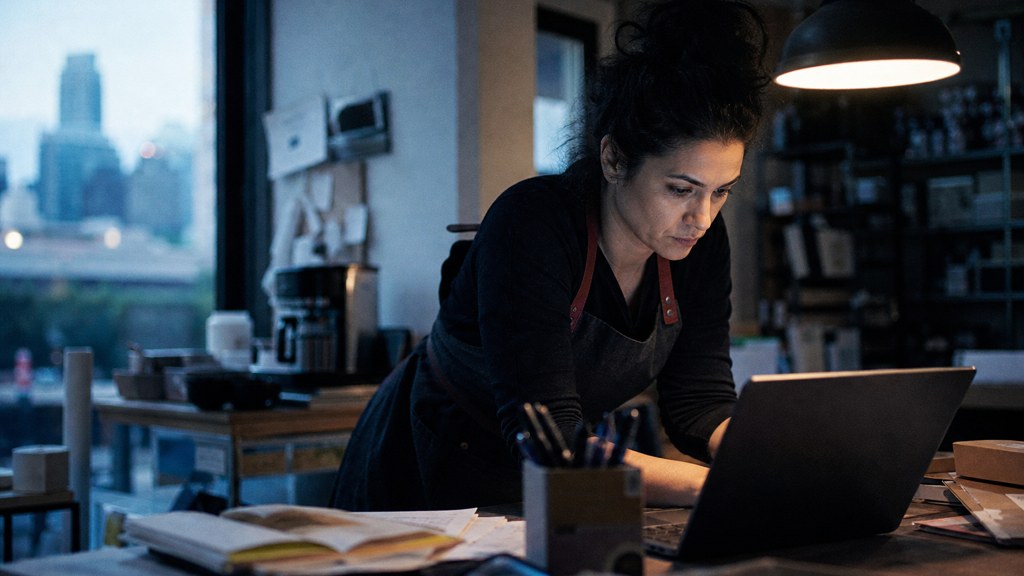 A small-business owner in an apron leaning over a laptop on a cluttered back-office desk late in the day, photographed in Austin