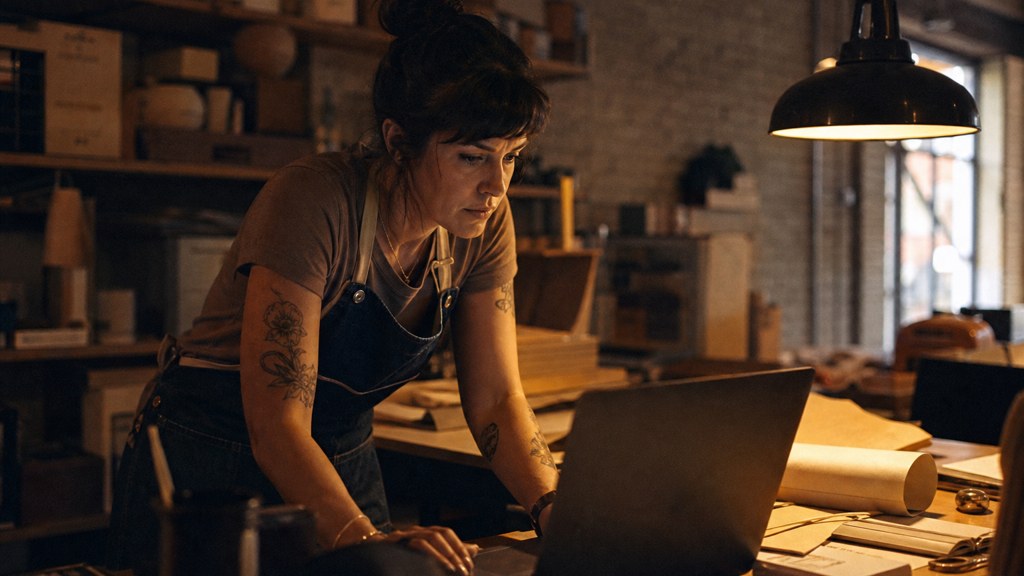 A small-business owner in an apron leaning over a laptop on a cluttered back-office desk late in the day, photographed in Austin