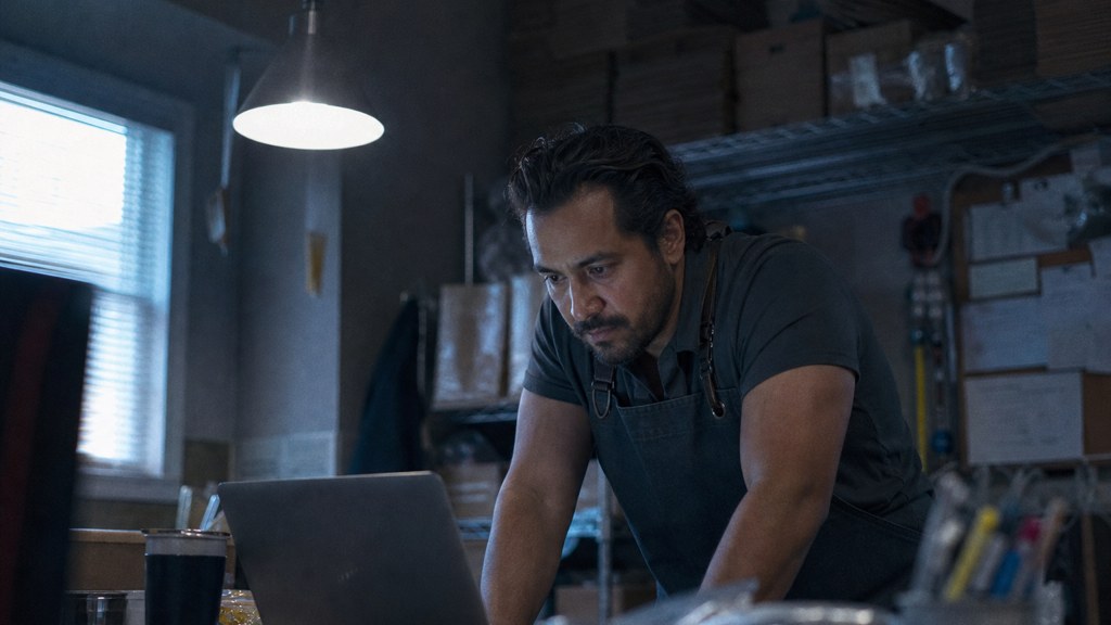A small-business owner in an apron leaning over a laptop on a cluttered back-office desk late in the day, photographed in Bakersfield