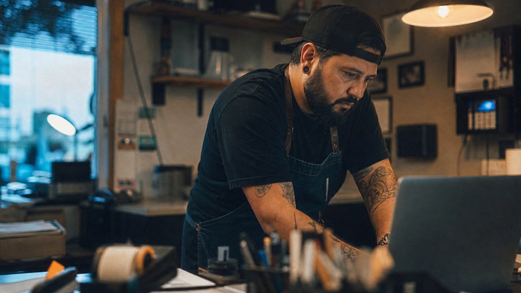 A small-business owner in an apron leaning over a laptop on a cluttered back-office desk late in the day, photographed in Bakersfield