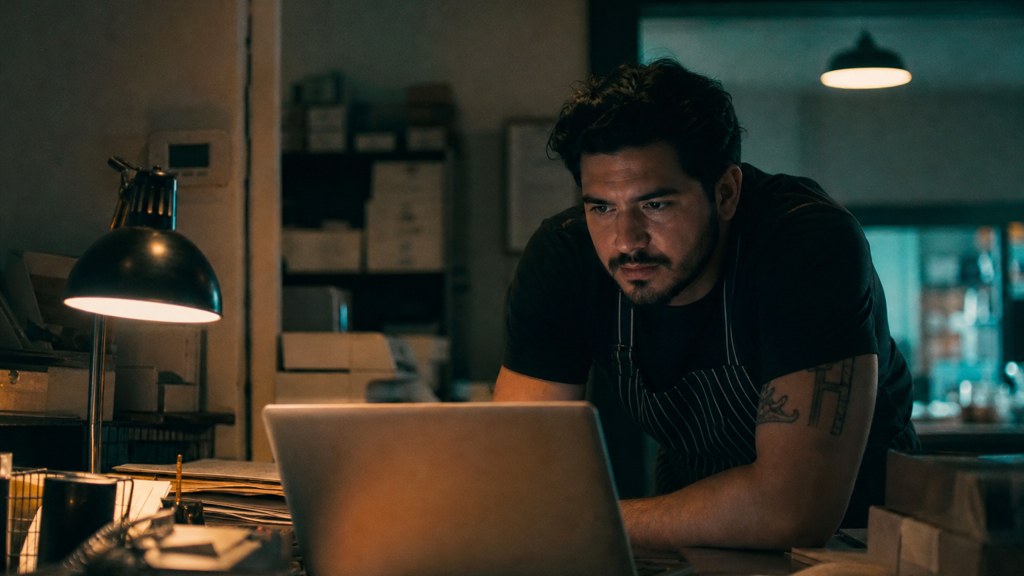 A small-business owner in an apron leaning over a laptop on a cluttered back-office desk late in the day, photographed in Bakersfield
