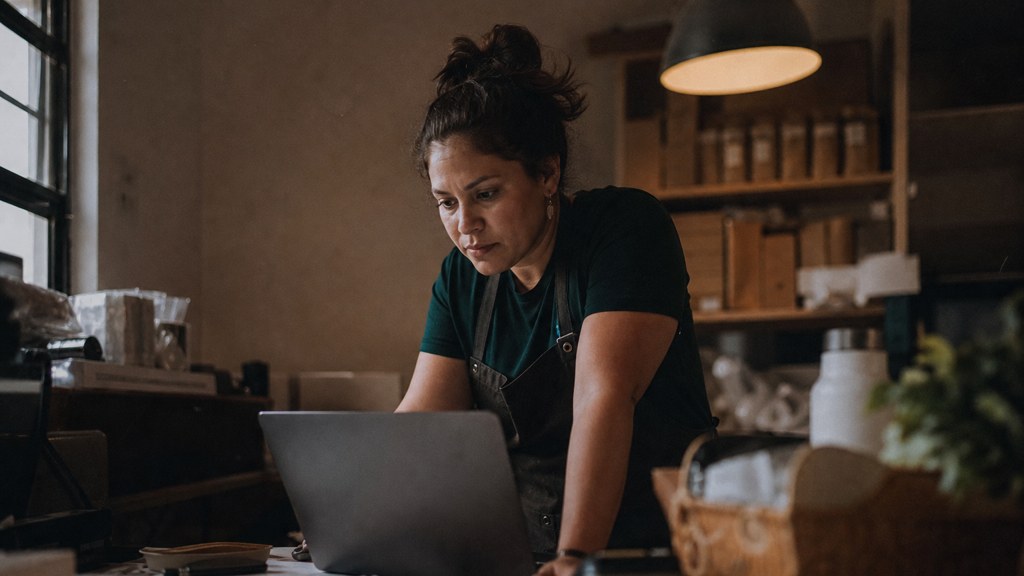 A small-business owner in an apron leaning over a laptop on a cluttered back-office desk late in the day, photographed in Bakersfield
