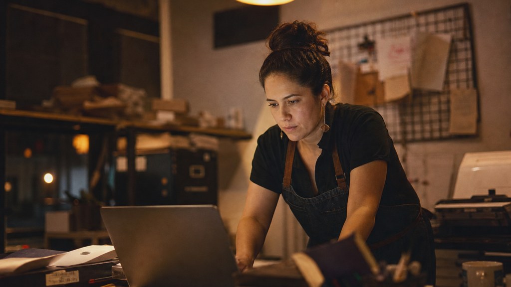 A small-business owner in an apron leaning over a laptop on a cluttered back-office desk late in the day, photographed in Bakersfield