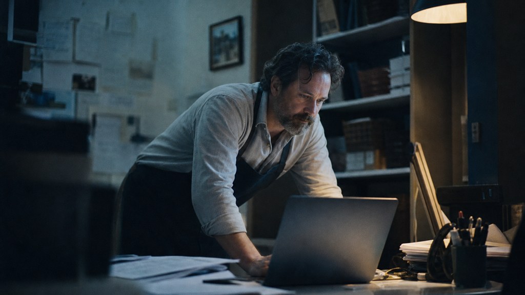 A small-business owner in an apron leaning over a laptop on a cluttered back-office desk late in the day, photographed in Baton Rouge