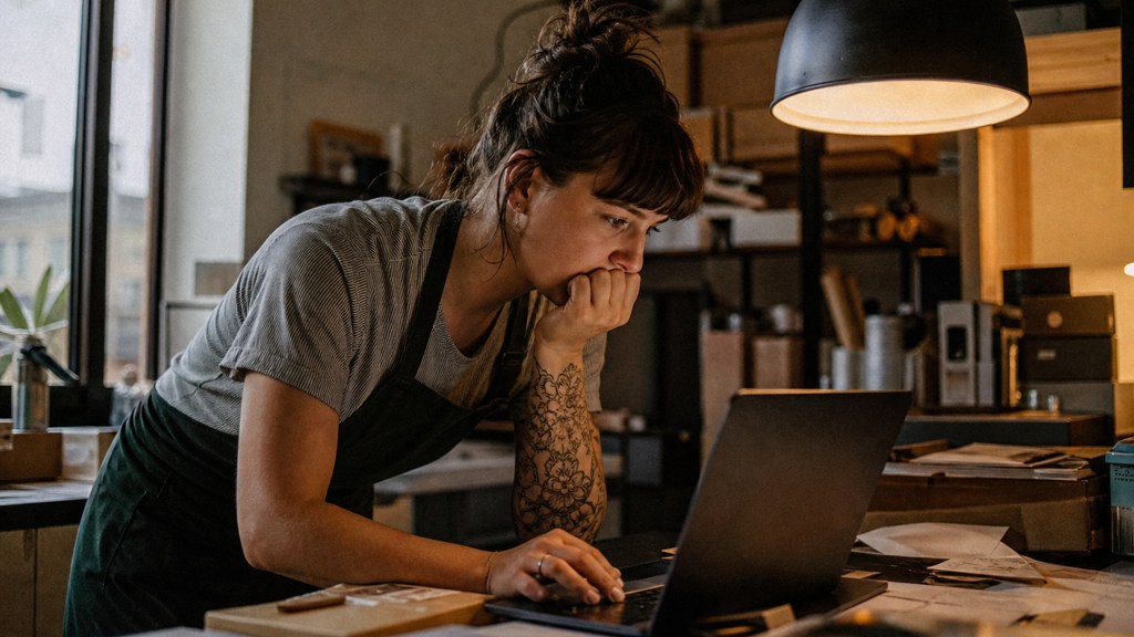 A small-business owner in an apron leaning over a laptop on a cluttered back-office desk late in the day, photographed in Baton Rouge