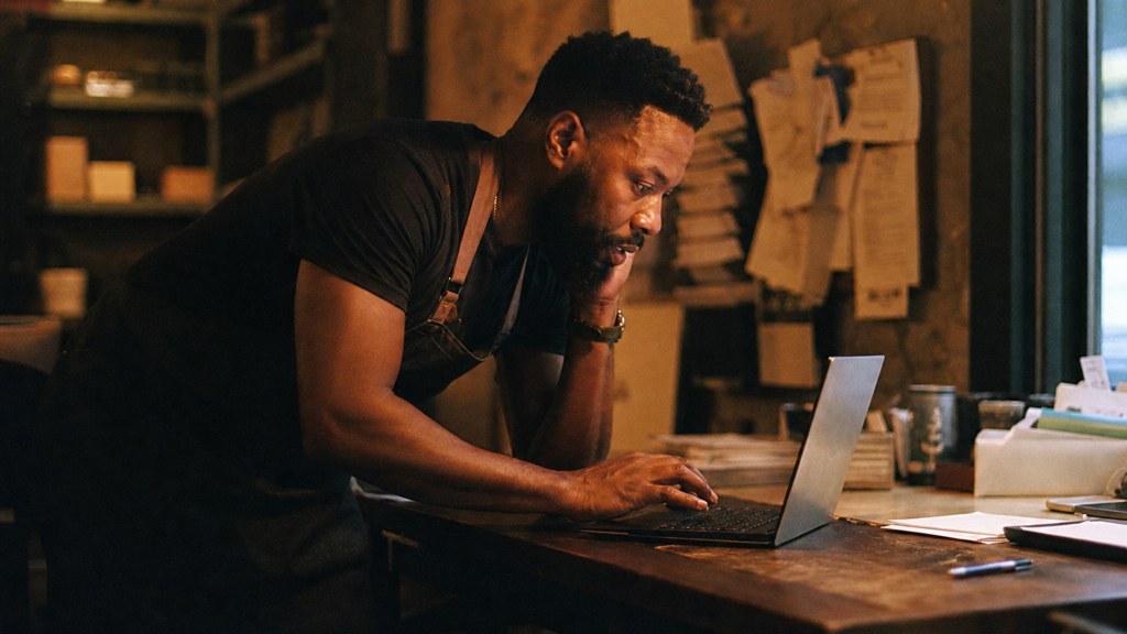 A small-business owner in an apron leaning over a laptop on a cluttered back-office desk late in the day, photographed in Baton Rouge