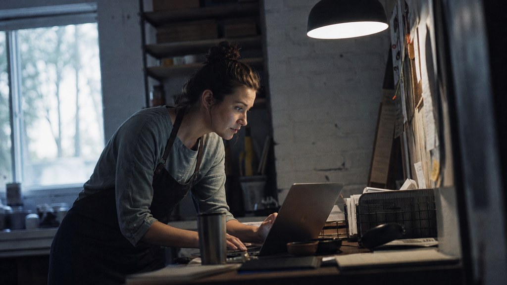A small-business owner in an apron leaning over a laptop on a cluttered back-office desk late in the day, photographed in Baton Rouge